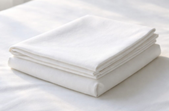 Folded Sleep and Beyond organic cotton percale pillowcase on a clean white bed in soft natural daylight