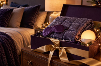 Open purple gift box on a nightstand holding a weighted blanket, silk sleep mask and glowing sunrise lamp